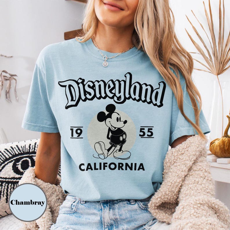 May include: Light blue t-shirt featuring the Disneyland logo in black script, with a graphic of Mickey Mouse and the text "1955 CALIFORNIA". The shirt has short sleeves and a crew neck.