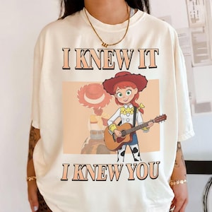 May include: An off-white t-shirt featuring a graphic of a cartoon cowgirl with red hair and a red hat playing an acoustic guitar. The text "I KNEW IT" is printed above the graphic, and "I KNEW YOU" is printed below in a bold, dark font.