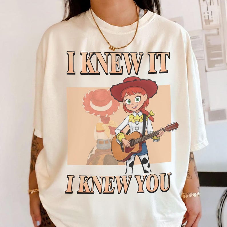 May include: An off-white t-shirt featuring a graphic of a cartoon cowgirl with red hair and a red hat playing an acoustic guitar. The text "I KNEW IT" is printed above the graphic, and "I KNEW YOU" is printed below in a bold, dark font.