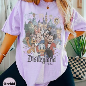 May include: A light purple t-shirt featuring a Disneyland graphic with Mickey Mouse, Minnie Mouse, Donald Duck, Goofy, and Pluto. The graphic includes a castle, fireworks, and a Ferris wheel. The text "Disneyland Est. 1955" is printed below the characters.