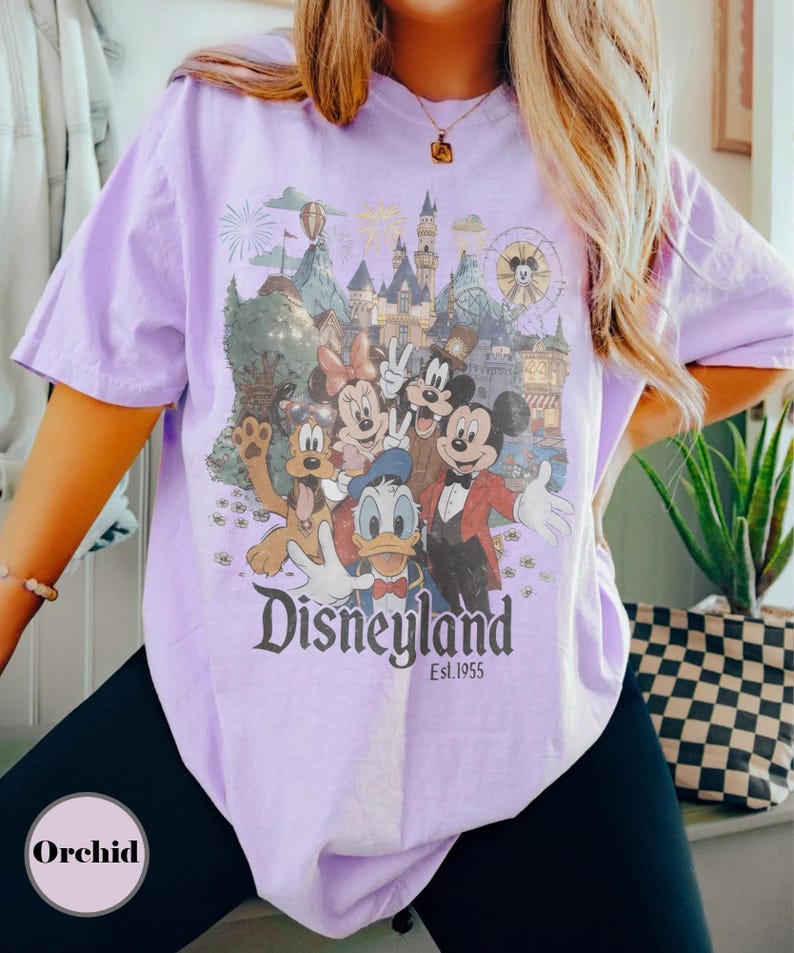 May include: A light purple t-shirt featuring a Disneyland graphic with Mickey Mouse, Minnie Mouse, Donald Duck, Goofy, and Pluto. The graphic includes a castle, fireworks, and a Ferris wheel. The text "Disneyland Est. 1955" is printed below the characters.