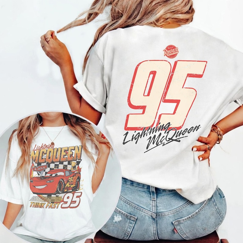 May include: White t-shirt featuring a graphic of Lightning McQueen and the number 95. The back of the shirt displays a large number 95 with the words "Lightning McQueen". The shirt is worn with blue jeans.