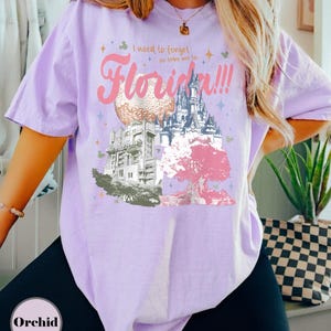 May include: A light purple t-shirt with a graphic design featuring the text "I need to forget so take me to Florida!!!" and illustrations of Disney World landmarks. The design includes a castle, a tree, and other park elements.