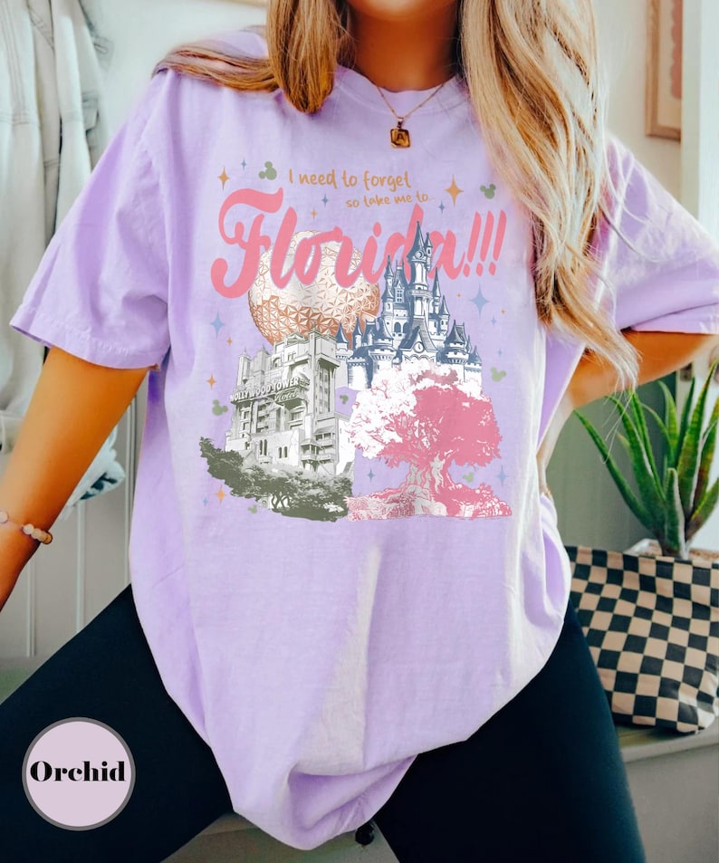 May include: A light purple t-shirt with a graphic design featuring the text "I need to forget so take me to Florida!!!" and illustrations of Disney World landmarks. The design includes a castle, a tree, and other park elements.