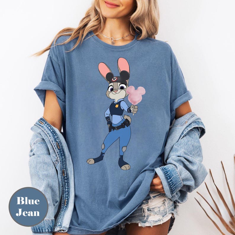 Puede incluir: Camiseta azul vaquero con una imagen de dibujos animados de Judy Hopps de Zootopia, sosteniendo un algod&oacute;n de az&uacute;car rosa. El personaje lleva un uniforme de polic&iacute;a. Las palabras "Blue Jean" est&aacute;n impresas en un c&iacute;rculo azul en la parte inferior izquierda.