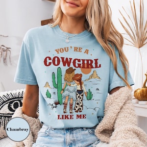 May include: A light blue short-sleeve t-shirt featuring a graphic print of two figures in cowboy hats standing in a desert landscape with cacti. The text reads YOU'RE A COWGIRL LIKE ME in red lettering. The shirt has a crew neckline and a relaxed fit.