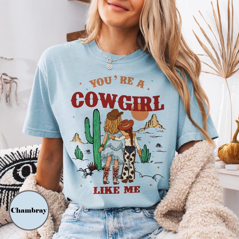 May include: A light blue short-sleeve t-shirt featuring a graphic print of two figures in cowboy hats standing in a desert landscape with cacti. The text reads YOU'RE A COWGIRL LIKE ME in red lettering. The shirt has a crew neckline and a relaxed fit.