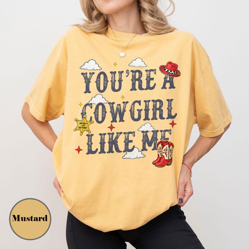 Puede incluir: Una camiseta de manga corta color amarillo mostaza con el texto "YOU'RE A COWGIRL LIKE ME" en letras azul oscuro. El dise&ntilde;o incluye ilustraciones de un sombrero de vaquero rojo, una estrella de sheriff amarilla, botas rojas y nubes blancas.