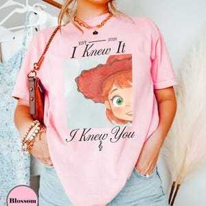 May include: A light pink short-sleeve t-shirt featuring a graphic print of a cartoon character with red hair and a red hat. The text reads EST 2026 I Knew It I Knew You with a small musical note symbol. The shirt has a crew neckline and a relaxed fit.