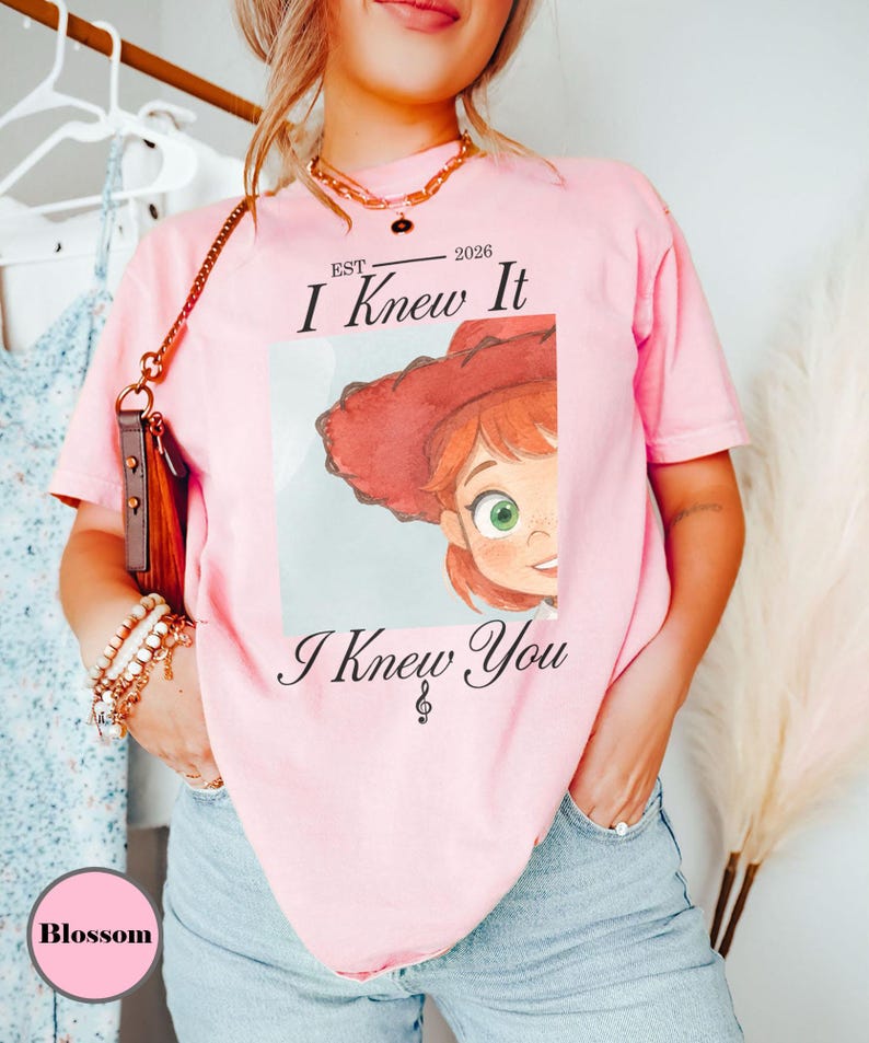 May include: A light pink short-sleeve t-shirt featuring a graphic print of a cartoon character with red hair and a red hat. The text reads EST 2026 I Knew It I Knew You with a small musical note symbol. The shirt has a crew neckline and a relaxed fit.