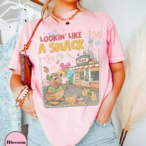 May include: Pink t-shirt with the text "Lookin' Like A Snack" and a colorful graphic featuring a cartoon mouse, balloons, and a snack stand. The design includes fireworks and a castle. The shirt is a light pink color.