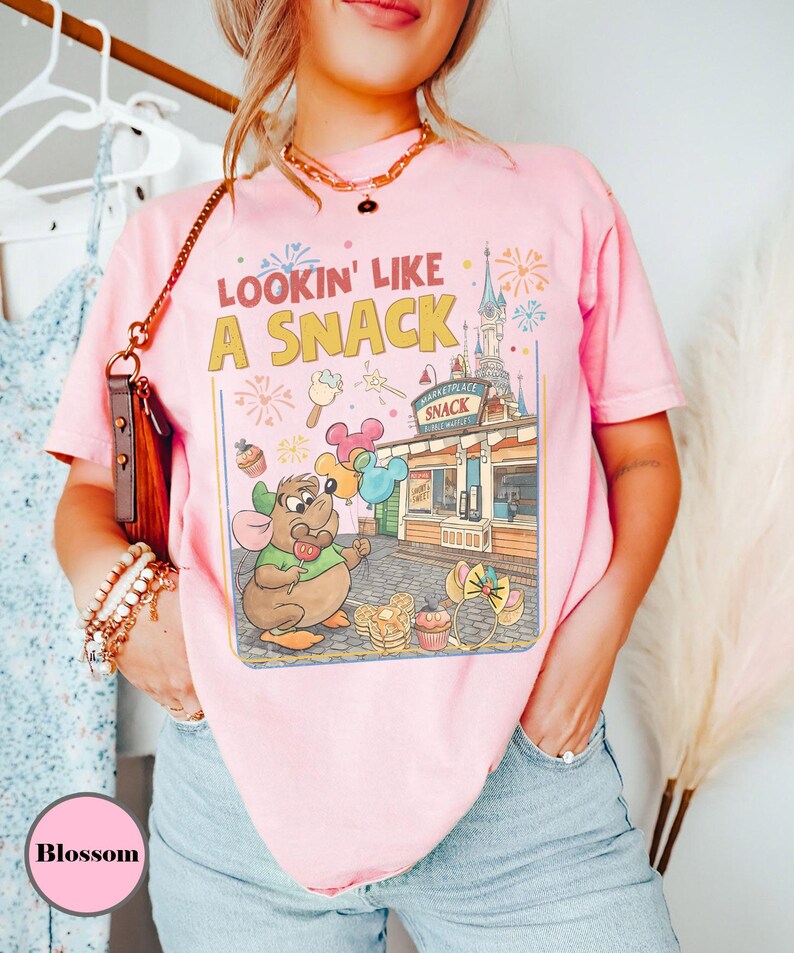 May include: Pink t-shirt with the text "Lookin' Like A Snack" and a colorful graphic featuring a cartoon mouse, balloons, and a snack stand. The design includes fireworks and a castle. The shirt is a light pink color.