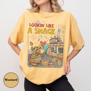 May include: Mustard-colored t-shirt with the phrase "Lookin' Like A Snack" and a cartoon illustration of a mouse holding a lollipop and balloons. The graphic includes a snack stand and a castle.