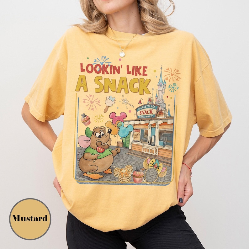 May include: Mustard-colored t-shirt with the phrase "Lookin' Like A Snack" and a cartoon illustration of a mouse holding a lollipop and balloons. The graphic includes a snack stand and a castle.