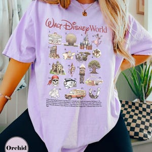 May include: A lilac t-shirt with the text "Walt Disney World" in a cursive font. The shirt displays illustrations of Disney attractions and characters, such as a carousel, Spaceship Earth, and a dragon. The word "Orchid" is printed at the bottom of the shirt.