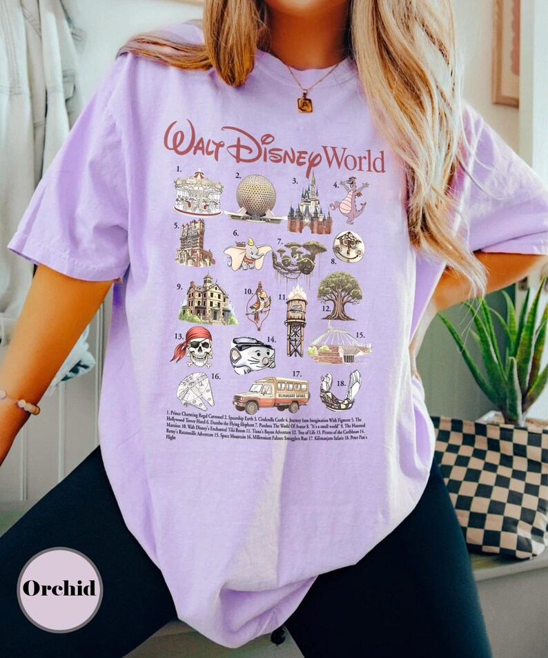 May include: A lilac t-shirt with the text "Walt Disney World" in a cursive font. The shirt displays illustrations of Disney attractions and characters, such as a carousel, Spaceship Earth, and a dragon. The word "Orchid" is printed at the bottom of the shirt.