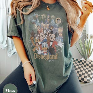 May include: Moss green t-shirt featuring a Disneyland graphic with Mickey Mouse, Minnie Mouse, Donald Duck, and other characters. The graphic includes a castle, fireworks, and the text "Disneyland Est.1955".