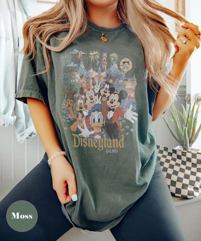 May include: Moss green t-shirt featuring a Disneyland graphic with Mickey Mouse, Minnie Mouse, Donald Duck, and other characters. The graphic includes a castle, fireworks, and the text "Disneyland Est.1955".