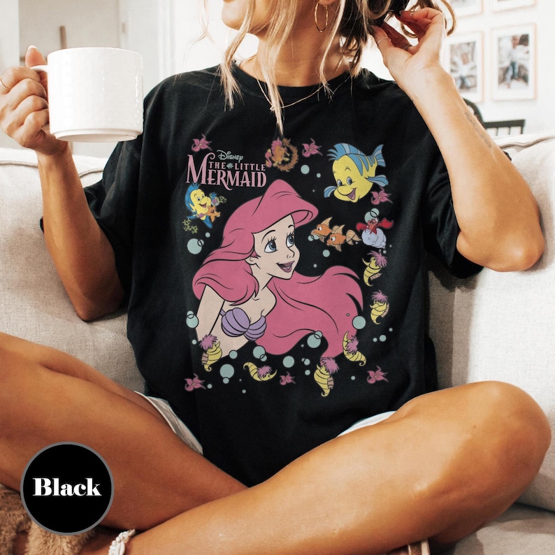 Puede incluir: Camiseta negra con un gr&aacute;fico de Ariel de La Sirenita de Disney. El dise&ntilde;o incluye a Ariel con pelo rosa, Flounder y otras criaturas marinas. Las palabras "Disney The Little Mermaid" est&aacute;n impresas encima de Ariel.