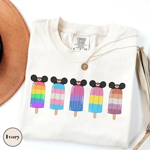 May include: Ivory-colored t-shirt featuring five colorful popsicle designs with black mouse ears. Each popsicle has a different color scheme, including rainbow, blue and pink, purple, yellow and pink, and orange and purple.