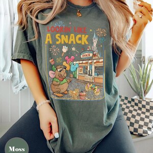 May include: Moss green t-shirt with a colorful graphic. The graphic features a cartoon mouse holding balloons in front of a snack shop with the text "Lookin' Like A Snack". Fireworks and ice cream are also in the design.