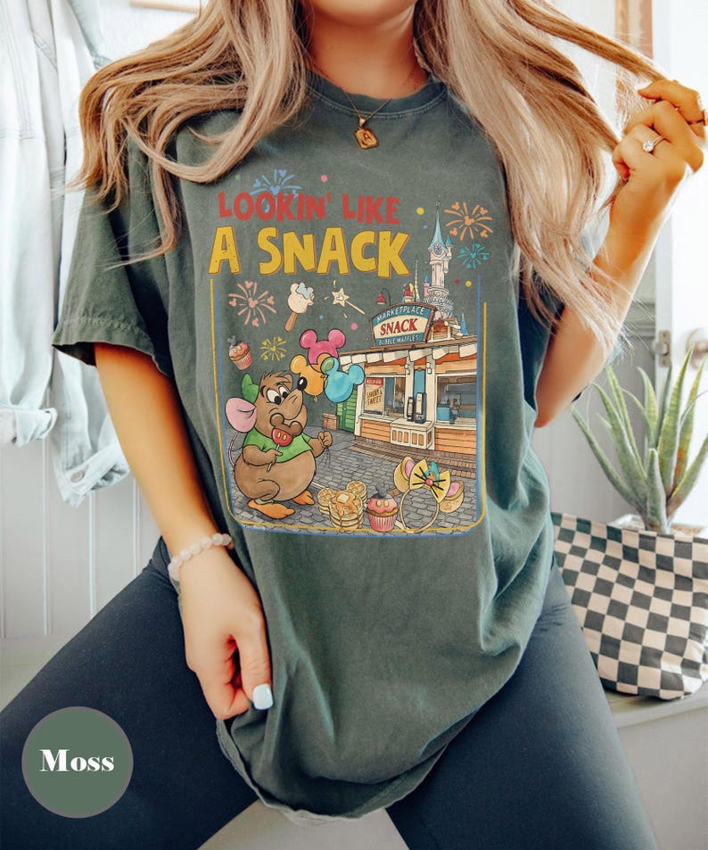 May include: Moss green t-shirt with a colorful graphic. The graphic features a cartoon mouse holding balloons in front of a snack shop with the text "Lookin' Like A Snack". Fireworks and ice cream are also in the design.