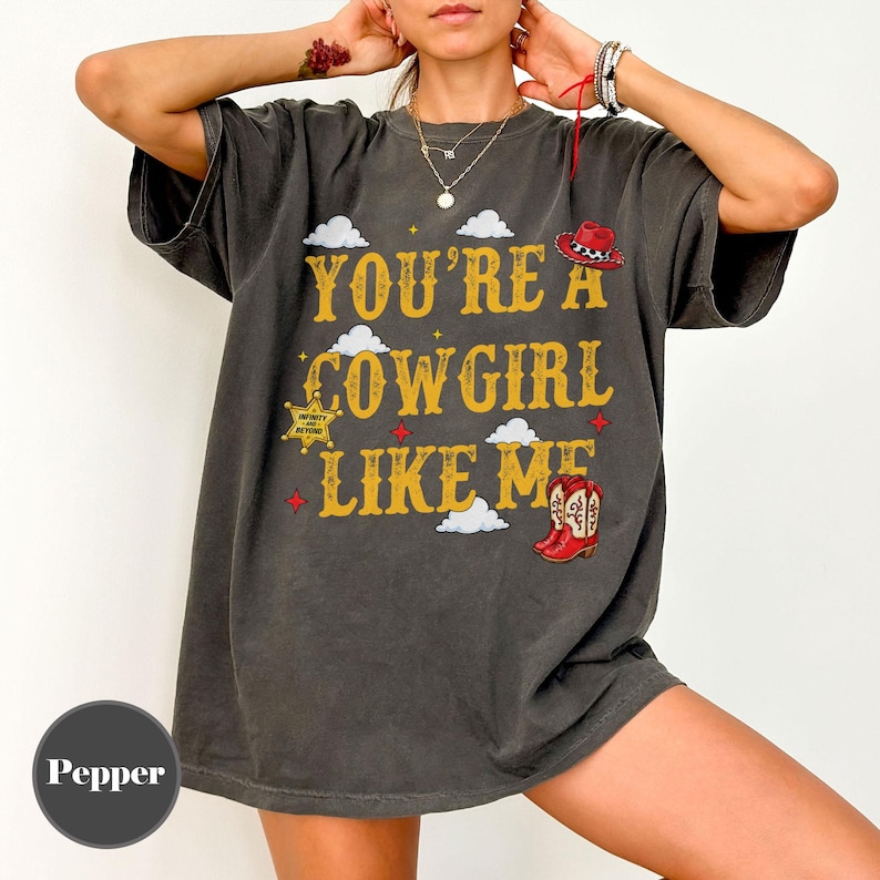 Puede incluir: Una camiseta gris carb&oacute;n de manga corta con el texto amarillo "YOU'RE A COWGIRL LIKE ME", con gr&aacute;ficos de un sombrero de vaquero rojo, una estrella de sheriff amarilla y botas de vaquero rojas, junto a nubes blancas y estrellas rojas.