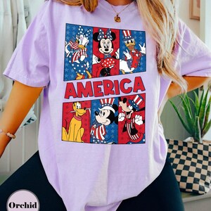 May include: A light purple t-shirt featuring a graphic of Disney characters in patriotic attire. The word "AMERICA" is displayed in red block letters. The design includes Donald Duck, Minnie Mouse, Mickey Mouse, and Goofy.