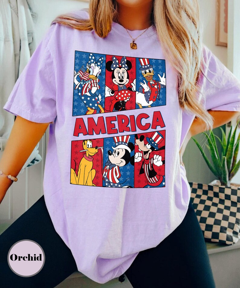 May include: A light purple t-shirt featuring a graphic of Disney characters in patriotic attire. The word "AMERICA" is displayed in red block letters. The design includes Donald Duck, Minnie Mouse, Mickey Mouse, and Goofy.