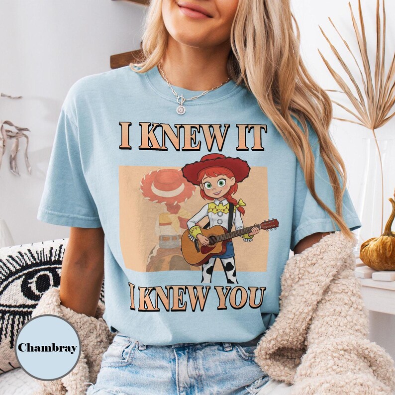 May include: A light blue t-shirt featuring a graphic of the character Jessie from Toy Story playing an acoustic guitar. The text "I KNEW IT" is printed above the character and "I KNEW YOU" is printed below in a bold, dark brown font.