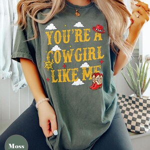 Puede incluir: Una camiseta de manga corta color verde musgo con el texto amarillo "YOU'RE A COWGIRL LIKE ME", decorada con gr&aacute;ficos de nubes blancas, un sombrero de vaquero rojo, una estrella de sheriff amarilla con "INFINITY AND BEYOND" y botas rojas.