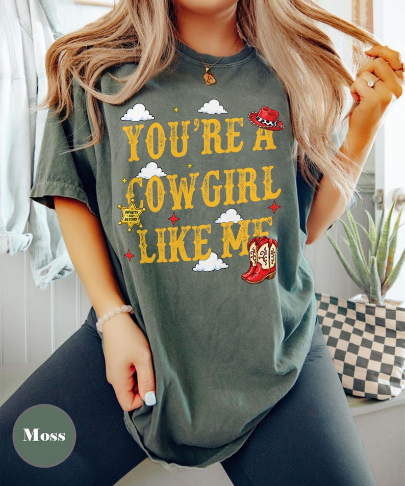 Puede incluir: Una camiseta de manga corta color verde musgo con el texto amarillo "YOU'RE A COWGIRL LIKE ME", decorada con gr&aacute;ficos de nubes blancas, un sombrero de vaquero rojo, una estrella de sheriff amarilla con "INFINITY AND BEYOND" y botas rojas.