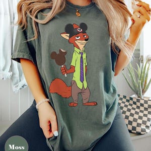 Puede incluir: Camiseta verde musgo con un personaje de zorro de dibujos animados que lleva un sombrero de Mickey Mouse y sostiene un helado con forma de Mickey Mouse. El zorro viste una camisa verde y una corbata morada. La camiseta es de corte holgado.