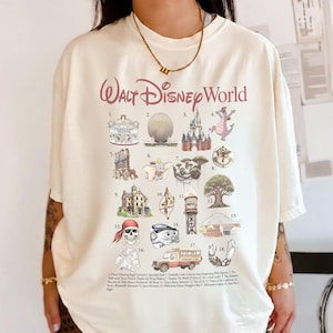 May include: Cream-coloured t-shirt with a graphic of Walt Disney World attractions. The design includes illustrations of iconic landmarks like Cinderella Castle, Spaceship Earth, and the Haunted Mansion, with the words "Walt Disney World" in a burgundy script.