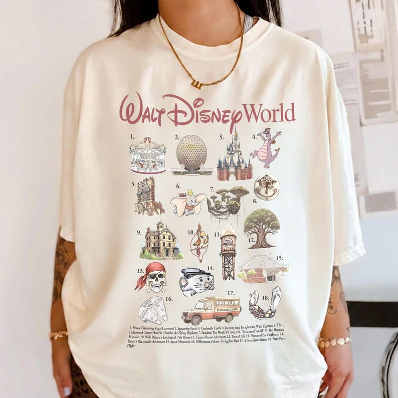 May include: Cream-coloured t-shirt with a graphic of Walt Disney World attractions. The design includes illustrations of iconic landmarks like Cinderella Castle, Spaceship Earth, and the Haunted Mansion, with the words "Walt Disney World" in a burgundy script.