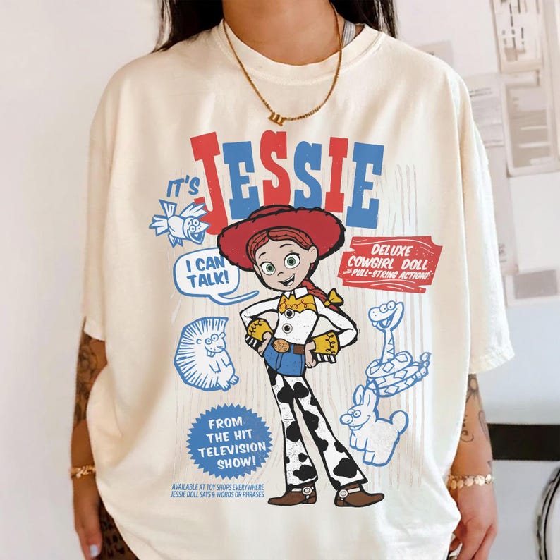 May include: Cream-colored t-shirt featuring a graphic of Jessie from Toy Story. The design includes the word "JESSIE" in red and blue, along with cartoon illustrations and text that reads "I CAN TALK!" and "DELUXE COWGIRL DOLL".