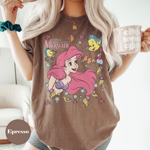Puede incluir: Camiseta color espresso con un gr&aacute;fico de Ariel de La Sirenita de Disney. El dise&ntilde;o incluye a Ariel con cabello rosa, un sujetador de concha p&uacute;rpura y varias criaturas marinas. La camiseta tiene un aspecto vintage y descolorido.