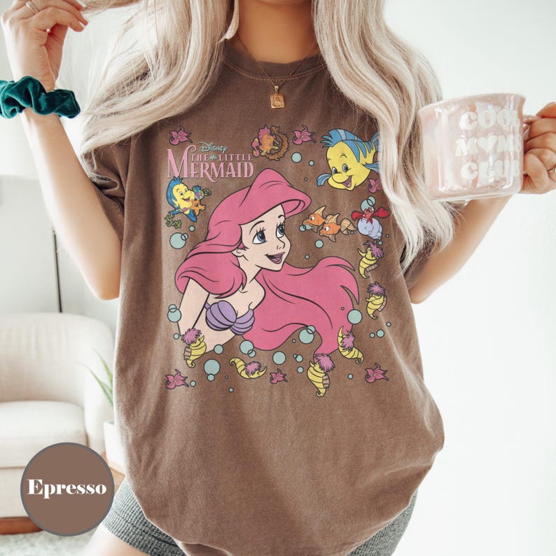 Puede incluir: Camiseta color espresso con un gr&aacute;fico de Ariel de La Sirenita de Disney. El dise&ntilde;o incluye a Ariel con cabello rosa, un sujetador de concha p&uacute;rpura y varias criaturas marinas. La camiseta tiene un aspecto vintage y descolorido.