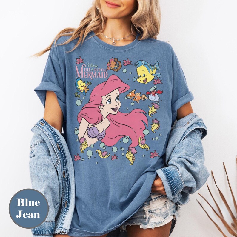 Puede incluir: Camiseta azul vaquero con un gr&aacute;fico de Ariel de La Sirenita de Disney. El dise&ntilde;o incluye a Ariel con pelo rosa, criaturas marinas y el t&iacute;tulo de la pel&iacute;cula. La camiseta es de color azul claro.