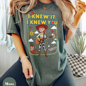 May include: A moss green t-shirt featuring a graphic of the character Jessie from Toy Story playing an acoustic guitar. The text reads I KNEW IT, I KNEW YOU. Surrounding elements include a sheriff badge, red cowboy boots, clouds, and stars.