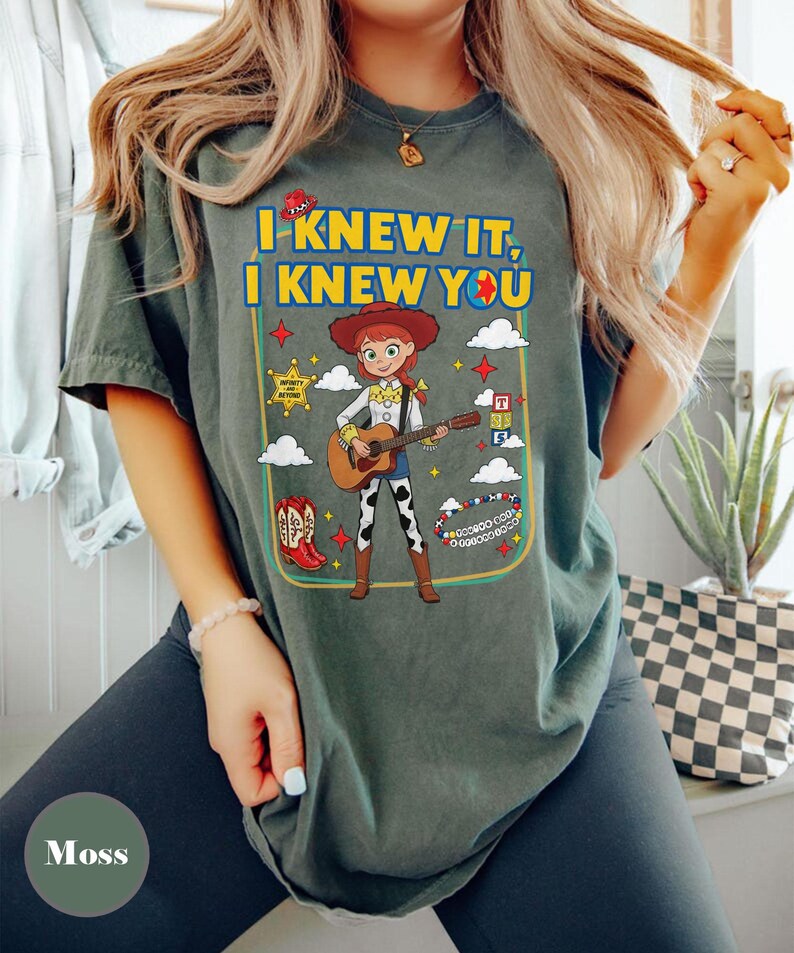 May include: A moss green t-shirt featuring a graphic of the character Jessie from Toy Story playing an acoustic guitar. The text reads I KNEW IT, I KNEW YOU. Surrounding elements include a sheriff badge, red cowboy boots, clouds, and stars.