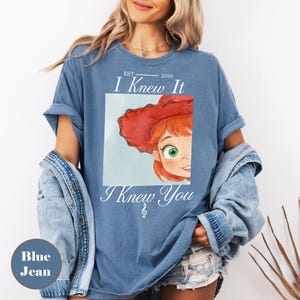 May include: A blue t-shirt featuring a graphic of a cartoon character with red hair and a red hat. The text reads "EST. 2026 I Knew It I Knew You" with a small musical note symbol. The shirt has short sleeves and a crew neckline, worn with a denim jacket.