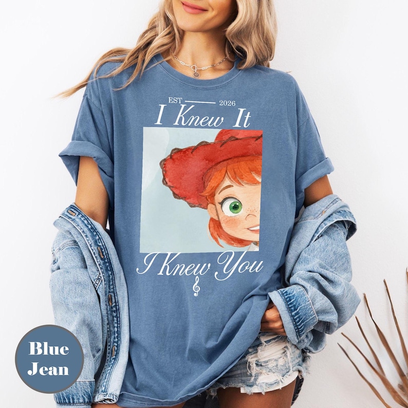 May include: A blue t-shirt featuring a graphic of a cartoon character with red hair and a red hat. The text reads "EST. 2026 I Knew It I Knew You" with a small musical note symbol. The shirt has short sleeves and a crew neckline, worn with a denim jacket.
