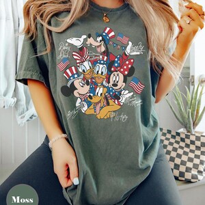 Peut inclure: Un t-shirt vert mousse avec un motif des personnages Disney Mickey Mouse, Minnie Mouse, Donald Duck, Daisy Duck, Dingo et Pluto, tous v&ecirc;tus de tenues patriotiques avec des drapeaux am&eacute;ricains. Le t-shirt porte les inscriptions "Mickey Mouse", "Minnie Mouse", "Daisy Duck", "Goofy" et "Pluto".