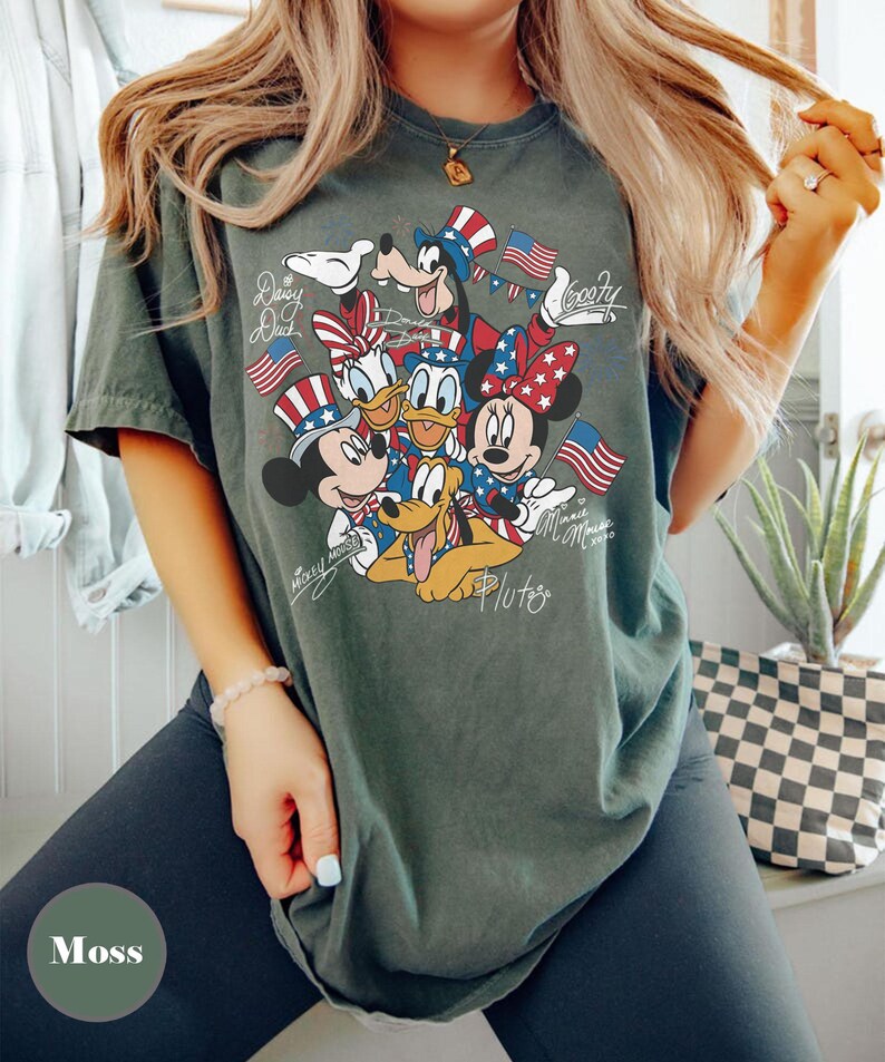 Peut inclure: Un t-shirt vert mousse avec un motif des personnages Disney Mickey Mouse, Minnie Mouse, Donald Duck, Daisy Duck, Dingo et Pluto, tous v&ecirc;tus de tenues patriotiques avec des drapeaux am&eacute;ricains. Le t-shirt porte les inscriptions "Mickey Mouse", "Minnie Mouse", "Daisy Duck", "Goofy" et "Pluto".