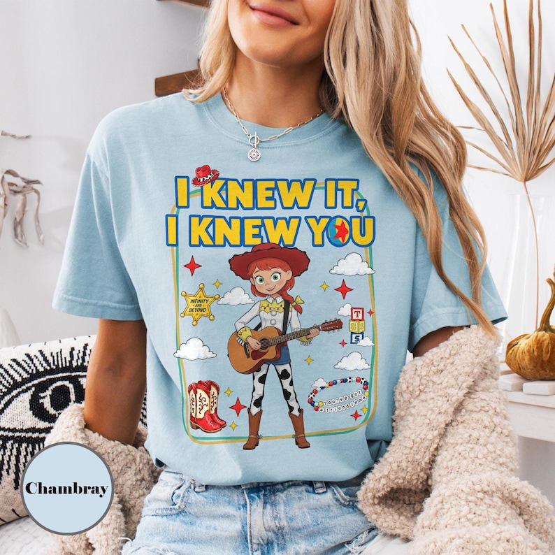 May include: A light blue t-shirt featuring a graphic of a cowgirl character playing an acoustic guitar. The text reads I KNEW IT, I KNEW YOU. The design includes a sheriff star, red boots, clouds, and colorful stars, all enclosed within a rectangular border.