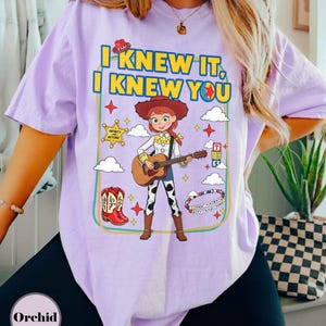May include: A light purple short-sleeve t-shirt featuring a graphic of a cowgirl character playing an acoustic guitar. The design includes the text "I KNEW IT, I KNEW YOU," a sheriff badge, red cowboy boots, alphabet blocks, and colorful beads.