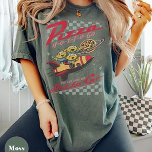 May include: Moss green t-shirt with a graphic featuring the text "Pizza H-O-T-T-O-Go" in red. The design includes cartoon aliens on a rocket ship with a pizza planet. The shirt has a vintage, distressed appearance.