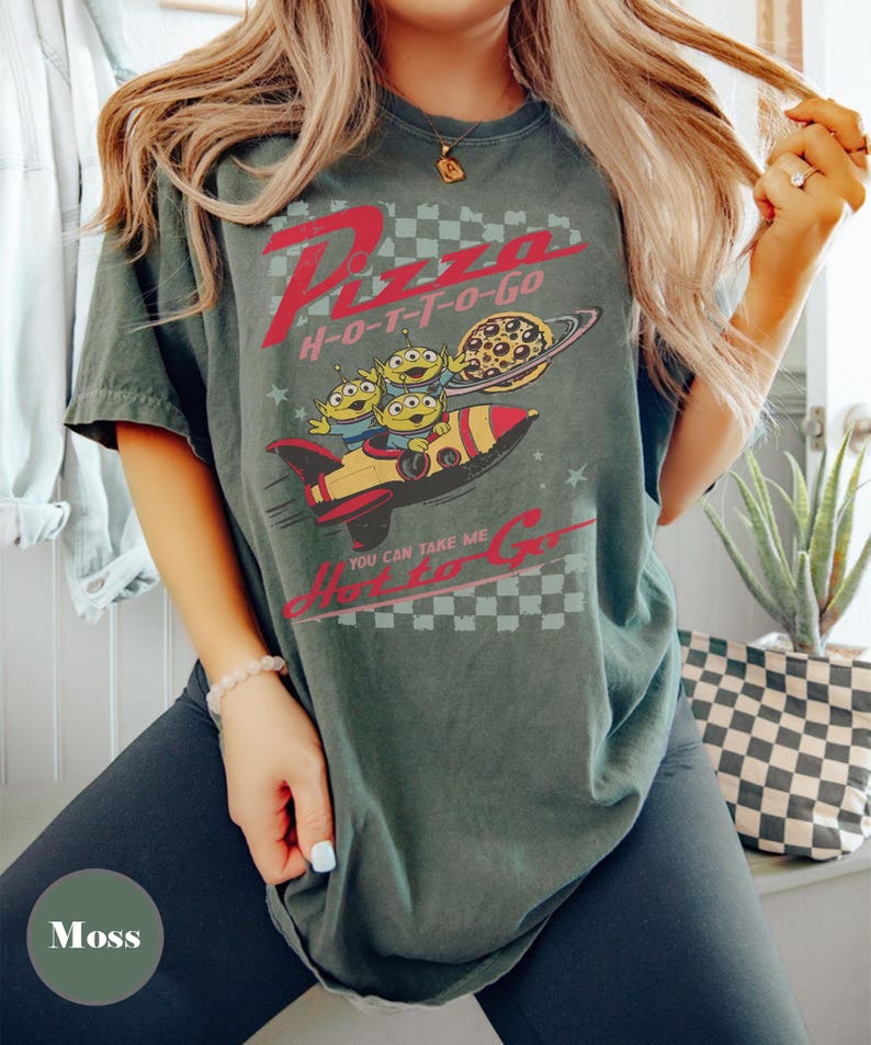 May include: Moss green t-shirt with a graphic featuring the text "Pizza H-O-T-T-O-Go" in red. The design includes cartoon aliens on a rocket ship with a pizza planet. The shirt has a vintage, distressed appearance.
