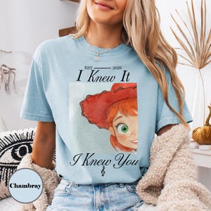 May include: A light blue short-sleeve t-shirt featuring a watercolor illustration of a character with red hair and a red hat. The text reads EST 2026 I Knew It I Knew You with a treble clef symbol. The shirt has a crew neckline and a relaxed fit.